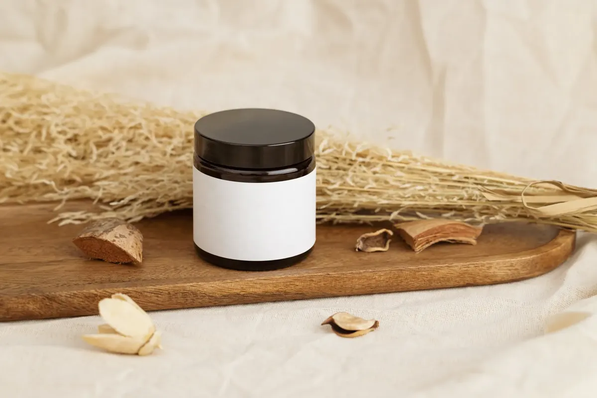 Natural cosmetic jar with blank label on a rustic wooden tray surrounded by dried herbs.